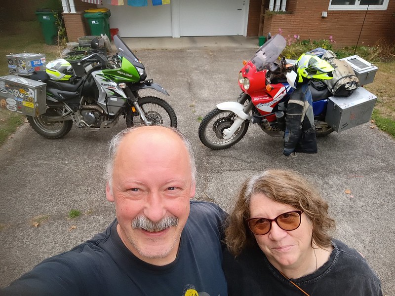 a man and a woman stand together
        taking a selfie with their motorcycles behind them